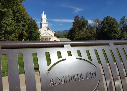 A bench with "Bountiful City" engraved, overlooking a church and trees against a clear blue sky.