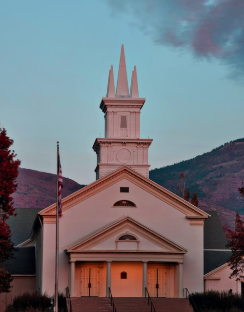 A white church with a tall spire, surrounded by trees, against a backdrop of mountains and a colorful sky at dusk.