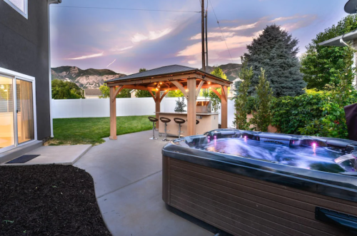 A backyard with a hot tub, gazebo, bar stools, and mountains in the background under a colorful sunset sky.