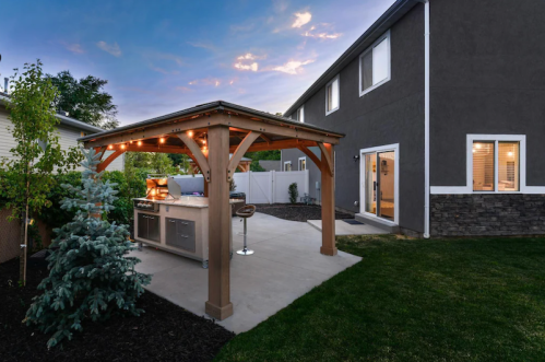 A backyard with a wooden gazebo featuring an outdoor kitchen, surrounded by grass and trees, under a twilight sky.