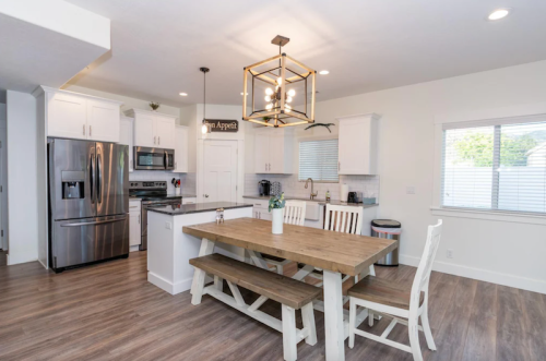Modern kitchen with white cabinets, stainless steel appliances, a wooden dining table, and large windows for natural light.