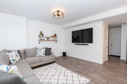 Modern living room with a gray sectional sofa, wall-mounted TV, and decorative shelf against a white wall.