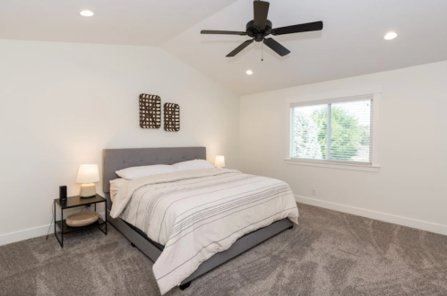 Cozy bedroom with a gray bed, two lamps, a ceiling fan, and a window overlooking greenery.