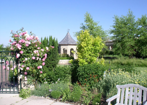 A sunny garden scene featuring blooming flowers, lush greenery, and a small stone gazebo in the background.