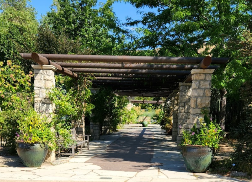 A stone archway covered with greenery leads to a garden path, flanked by large planters and benches.