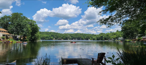 A serene lake scene with fluffy clouds, lush greenery, and a kayak on the water, framed by trees and a wooden dock.