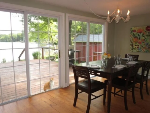 A dining area with a wooden table and chairs, featuring large windows overlooking a deck and a cat outside.