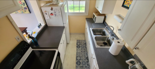 A bright kitchen with white cabinets, a black countertop, a sink, and appliances, featuring a patterned rug on the floor.