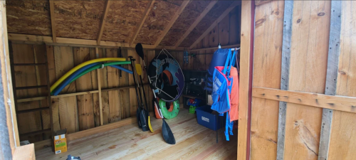 A wooden shed interior with paddles, a kayak, and colorful life jackets hanging on the wall.