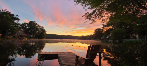 A serene lake at sunset, with colorful clouds reflecting on the water and a wooden dock with a chair in the foreground.