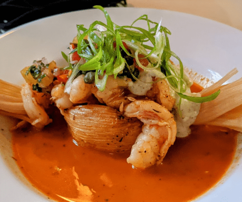 A plate of shrimp served on a tamale, topped with green onions and a creamy sauce, surrounded by a flavorful broth.