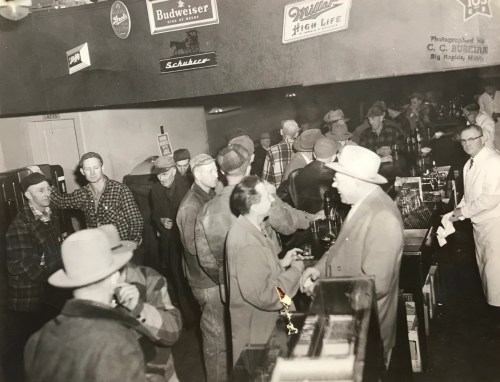 A crowded bar scene with men in hats chatting and drinking, vintage decor featuring beer signs on the walls.