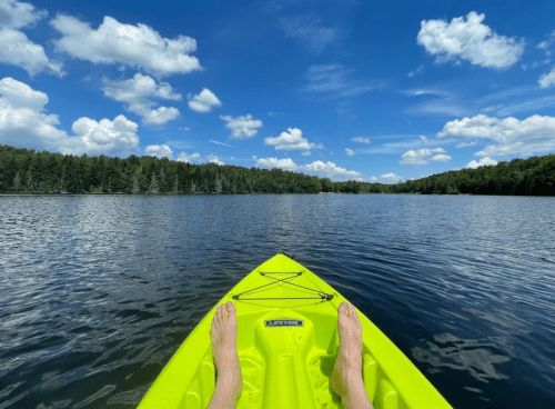 A view from a bright yellow kayak on calm water, with feet resting on the edge and a blue sky filled with clouds above.