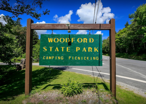 Sign for Woodford State Park, indicating camping and picnicking areas, with a scenic background of trees and blue sky.