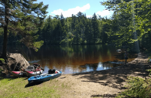A serene lake surrounded by trees, with kayaks on the shore and a person standing by the water's edge.