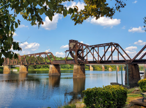 A rusty train bridge spans a calm river, surrounded by greenery and under a blue sky with fluffy clouds.