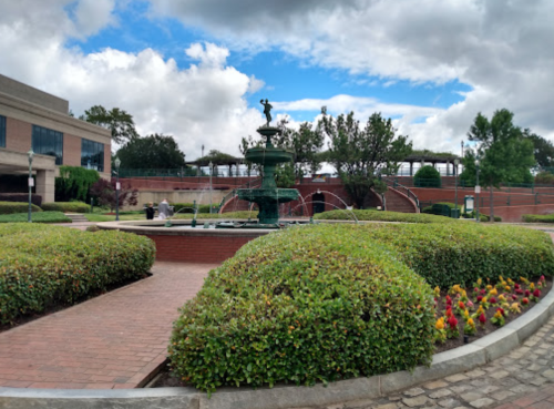 A green park with a fountain, surrounded by neatly trimmed bushes and colorful flowers under a partly cloudy sky.
