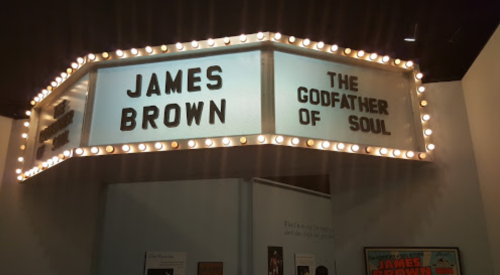 Sign featuring "James Brown" and "The Godfather of Soul" illuminated with lights in a museum setting.