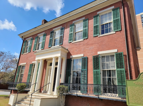 Historic brick building with green shutters, white columns, and a porch, set against a blue sky with clouds.