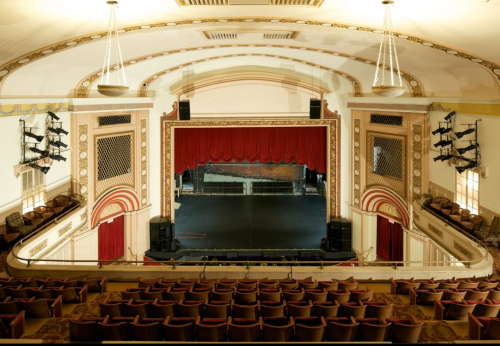 Interior of a vintage theater featuring a red curtain stage and ornate architectural details, with rows of seats visible.