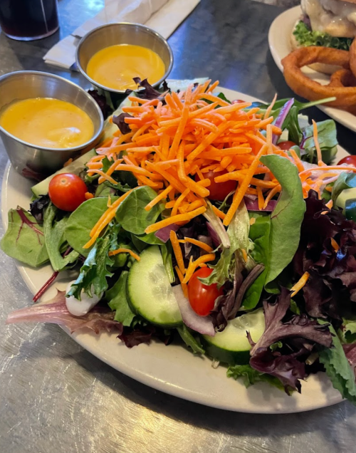 A fresh salad with mixed greens, cherry tomatoes, cucumber, and shredded carrots, served with two small cups of dressing.