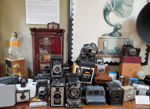 A collection of vintage cameras and photography equipment displayed on a table, with a gramophone illustration in the background.