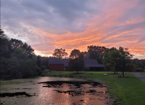 A serene pond reflects a vibrant sunset, with trees and a red barn in the background under a colorful sky.