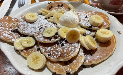 A plate of fluffy pancakes topped with banana slices, chocolate chips, and a scoop of ice cream, dusted with powdered sugar.