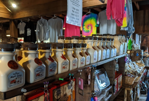 Bottles of Vermont maple syrup lined up on a wooden shelf in a rustic shop, with colorful clothing hanging above.