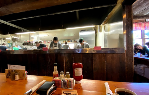 A cozy restaurant interior with a wooden table, condiments, and a view of chefs working in an open kitchen.