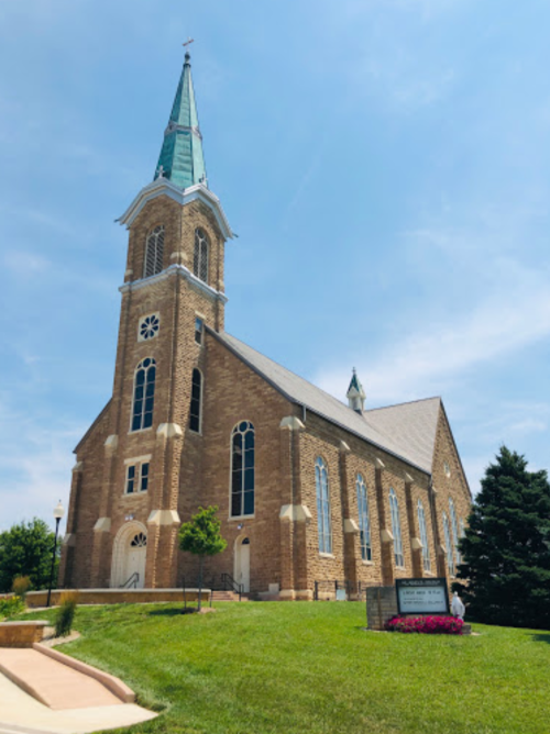 A tall stone church with a green spire, surrounded by grass and trees, under a clear blue sky.