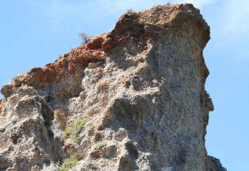 A rugged rock formation with a jagged peak against a clear blue sky, featuring sparse vegetation on its surface.