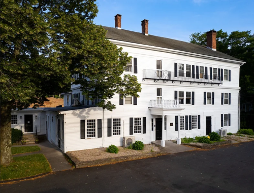 A three-story white building with black shutters, surrounded by trees and a paved area, under a clear blue sky.
