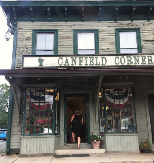 A green wooden building with "Canfield Corner" sign, featuring large windows and patriotic decorations.