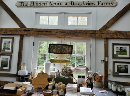 Interior of a farm shop with wooden beams, framed photos, and a sign reading "Sweet Corn" near various products.