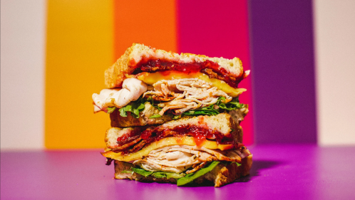 A stacked sandwich with layers of turkey, cheese, lettuce, and jam, set against a colorful striped background.
