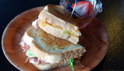A toasted sandwich with layers of ham, cheese, and lettuce, served on a wooden plate with a side of fruit.