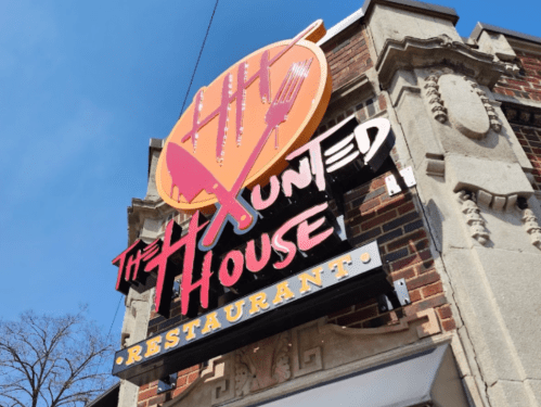 Sign for "The Haunted House Restaurant" featuring utensils and a playful design, mounted on a brick building.