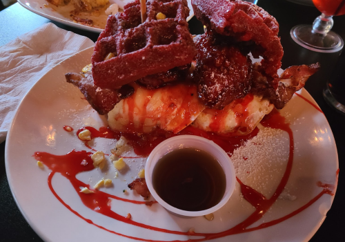 A plate featuring red velvet waffles, fried chicken, bacon, and syrup, drizzled with sauce and garnished with corn.