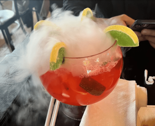 A vibrant red cocktail with lime slices and smoke, served in a large glass on a table.