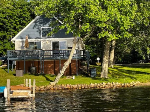 A lakeside house with a deck, surrounded by trees and grass, reflecting in the calm water.