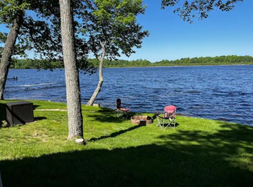 A serene lakeside view with green grass, two chairs, and a fire pit under clear blue skies and trees.