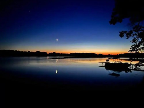 A serene lake at dusk, reflecting a deep blue sky with stars and a crescent moon, alongside a silhouette of a dock.