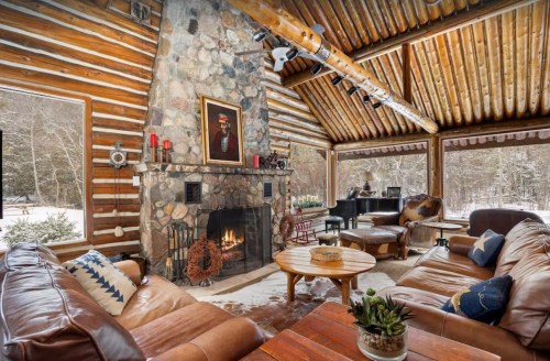 Cozy log cabin interior with a stone fireplace, wooden beams, and comfortable seating, surrounded by snowy scenery.