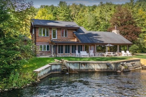 A rustic log cabin by a serene lake, surrounded by trees, with a porch and outdoor seating.