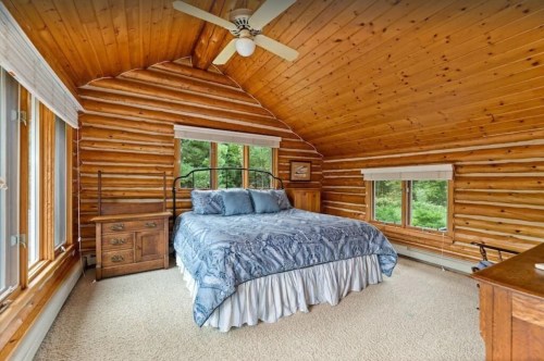Cozy log cabin bedroom with a queen bed, wooden furniture, and large windows overlooking greenery.