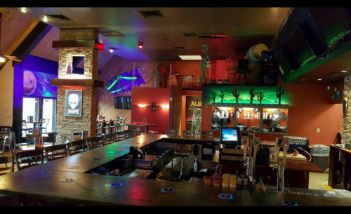 A vibrant bar interior with alien-themed decor, colorful lighting, and a stone counter.