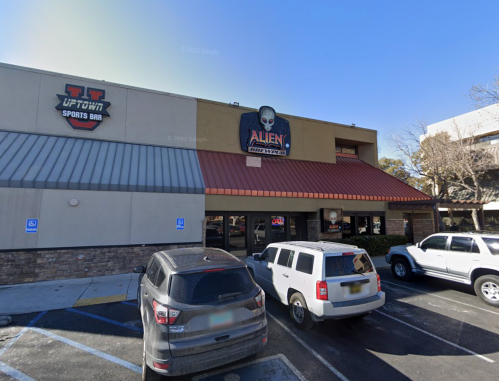 Exterior of a sports bar with a sign featuring an alien logo, surrounded by parked cars on a sunny day.