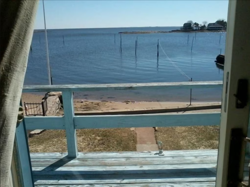 View from a porch overlooking a calm lake with sandy shore and distant houses on the opposite bank.