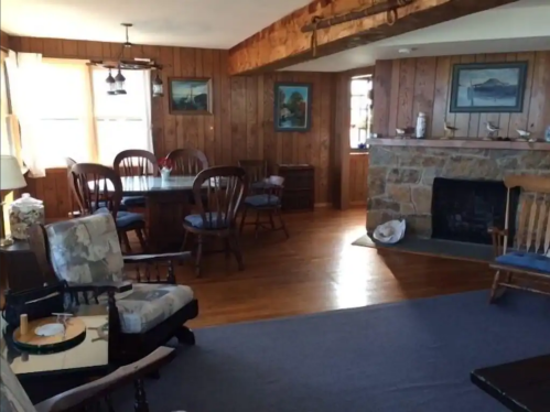 Cozy living room with wooden walls, a stone fireplace, dining area, and rustic furniture.